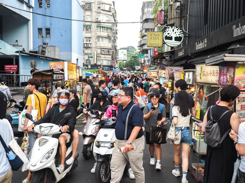 Looking up a crowded market street filled with people and some scooters squeezing through