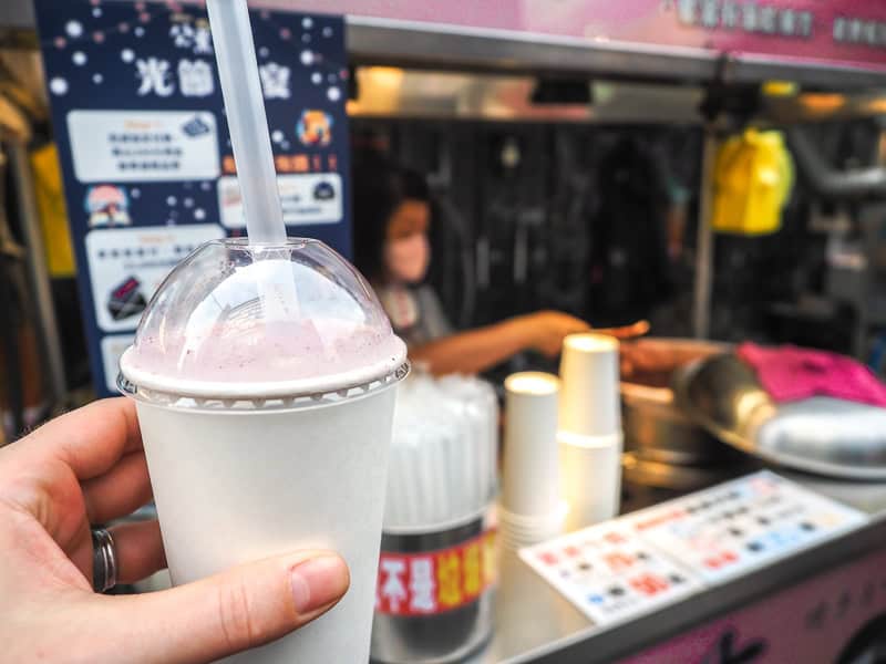 A hand holding up a white cup with plastic dome lid and purple drink inside on the left side, in front of the stall it was just purchased at