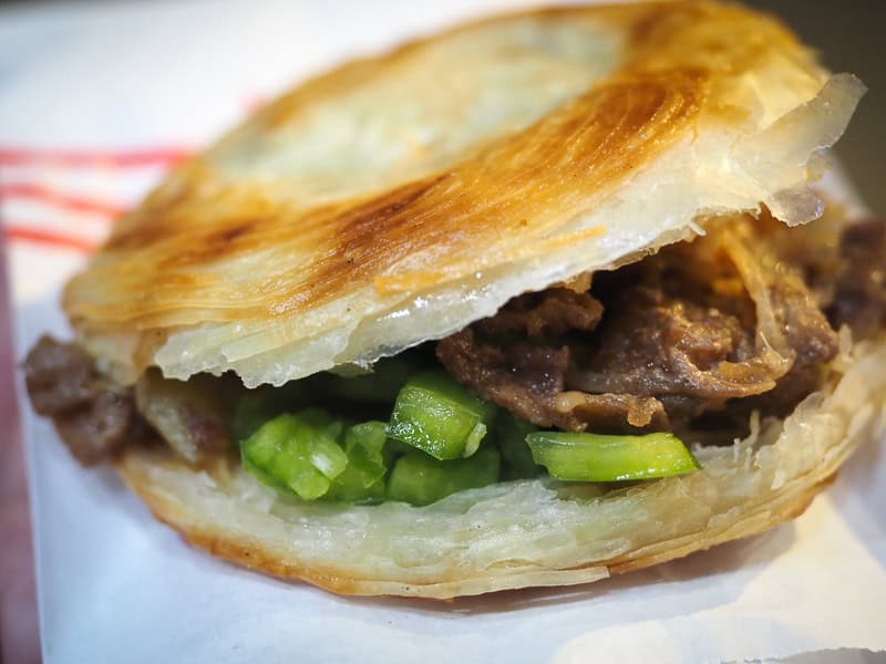 Close up of a pastry bun cut in half with pulled pork and green bell peppers inside on a white paper takeaway bag