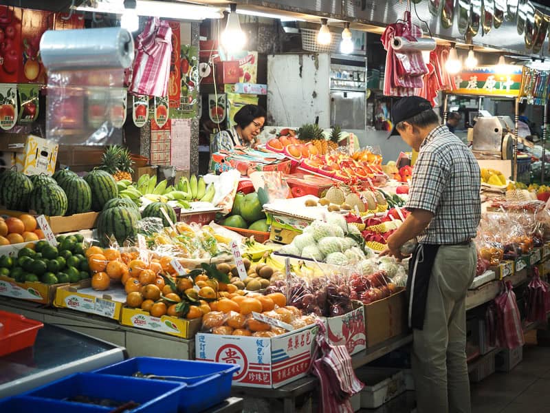 A fruit vendor in an indoor market, with one customer looking at the fruits