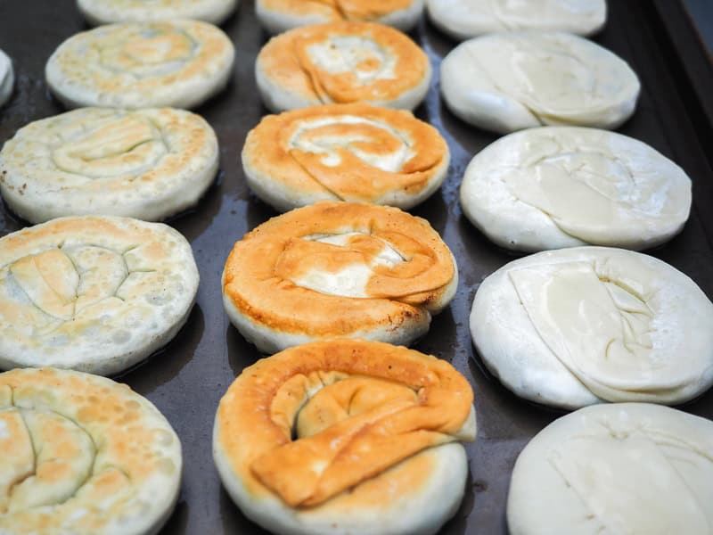 Looking up three rows of spiral green onion cakes on a flat grill, of varying levels of readiness to eat