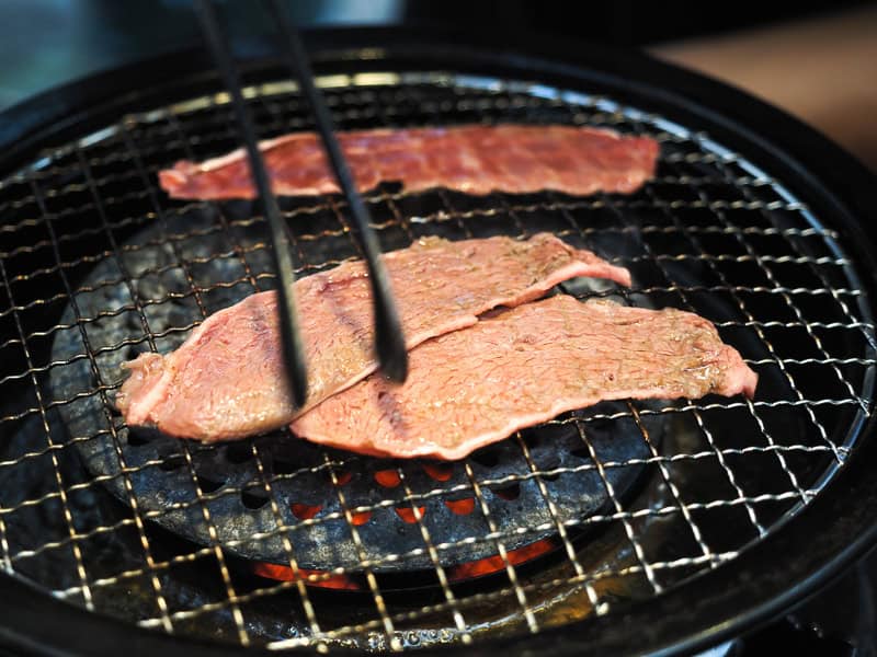 Close up of a small round grill with three strips of beef on it and tongs handling one of them