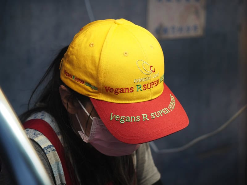A night market vendor tilting her head forward so we mostly only see her cap, which is yellow and red and has the words vegan on it