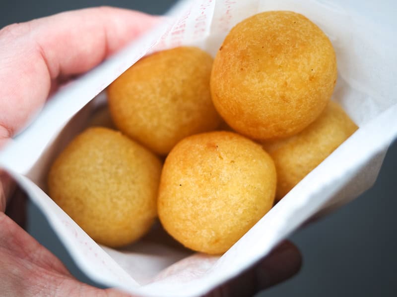 A hand holding a small white paper bag with 5 orange deep fried sweet potato balls inside it
