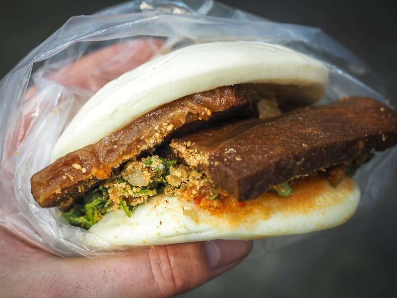 A hand holding a Taiwanese gua bao, or white steamed bun filled with two slabs of veggie meat, pickled greens, peanut powder, and spicy sauce