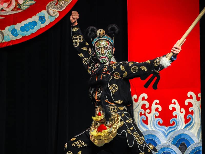A Taiwanese opera performer in mostly black costume holding out of a sword