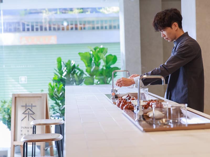 A man prepares tea on the right side of a long bar counter with shop window facing the street behind him