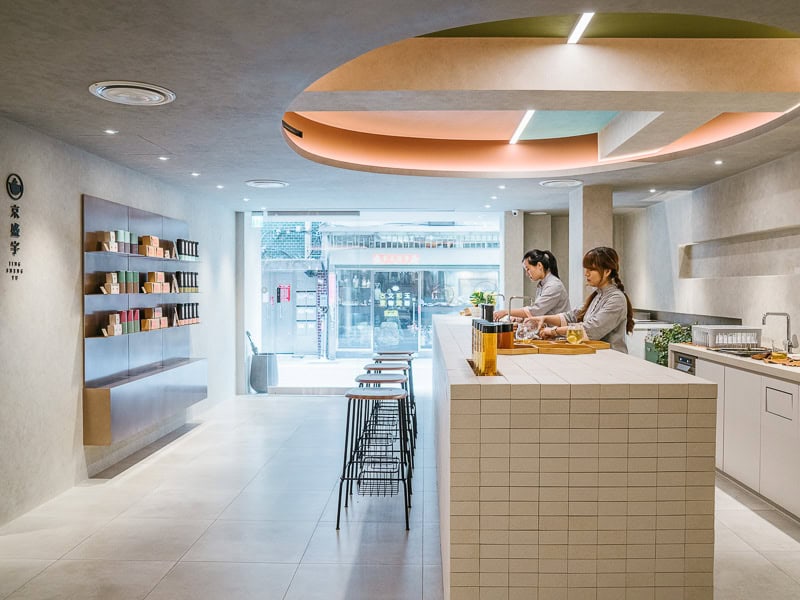 Looking at a very clean, modern looking tea shop from the back of the shop, with wall of products on the left, large bar with stools on the right, and two staff members behind the bar preparing tea