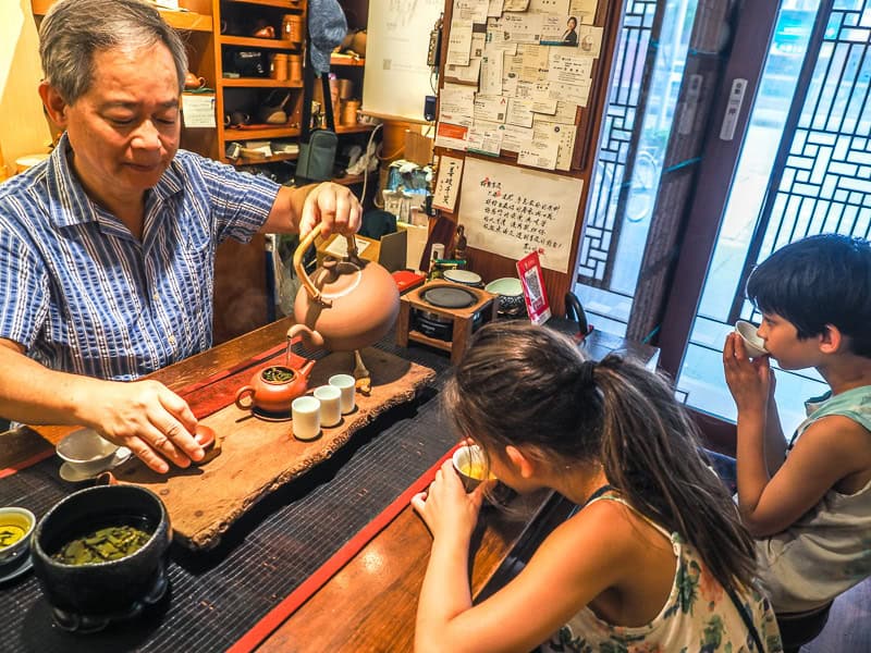 An elderly Taiwanese tea shop owner preparing tea in the traditional gongfu style with two kids seated across from him tasting the tea from little cups