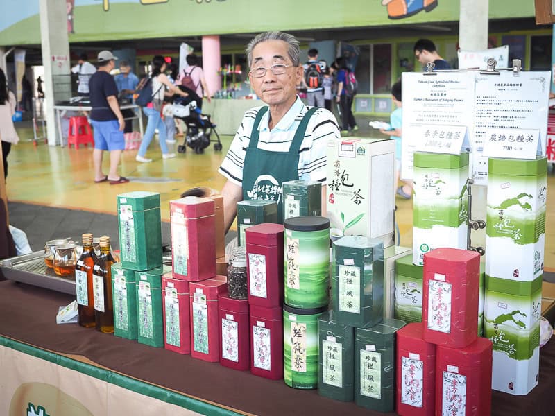 A vendor standing behind a table displaying boxes of tea at an outdoor farmer's market