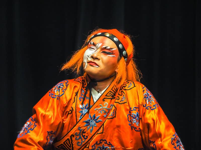Profile shot of a Taiwanese opera performer wearing mostly orange, with orange hair, black and white face paint, and a totally black background
