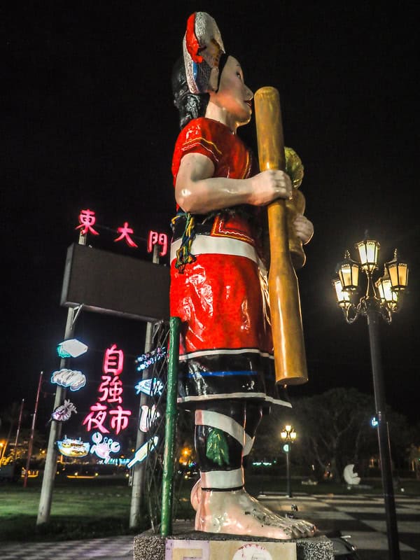 A tall statue of a female Taiwanese aboriginal in red costume holding a pestle, shot at night, next to a night market entrance