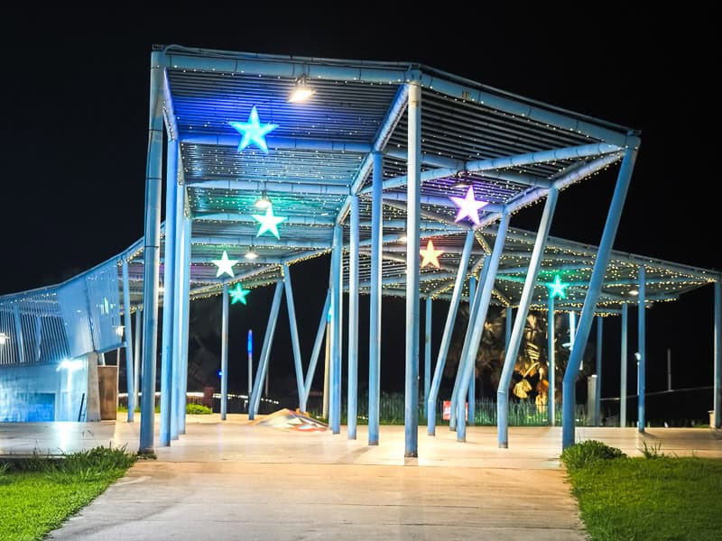 A park at night, with cement paths leading under two rows of metal beams with hanging lights and lit up stars