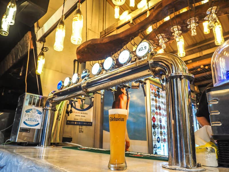 A pint of Eidelweiss beer on a bar with bar taps behind and hanging bottle lanterns above