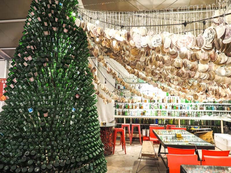 A tower of beer bottles that looks like a Christmas tree and sitting area beside it with hanging oyster shells above