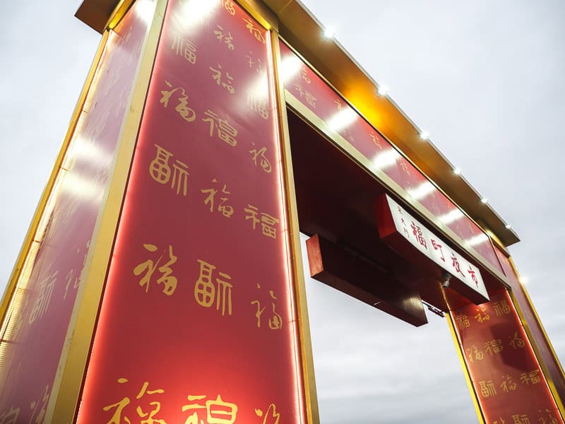 Looking up at a tall red and orange entrance gate to a night market from one side of it, with gray sky above