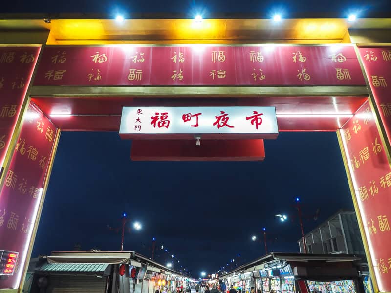Looking up a night market street at night through a tall red entrance gate with Chinese characters on it