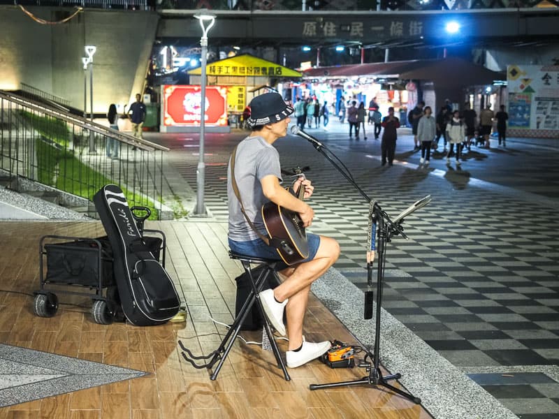 A Taiwanese man planning guitar and singing beside a night market, shot from the side and some people walk by