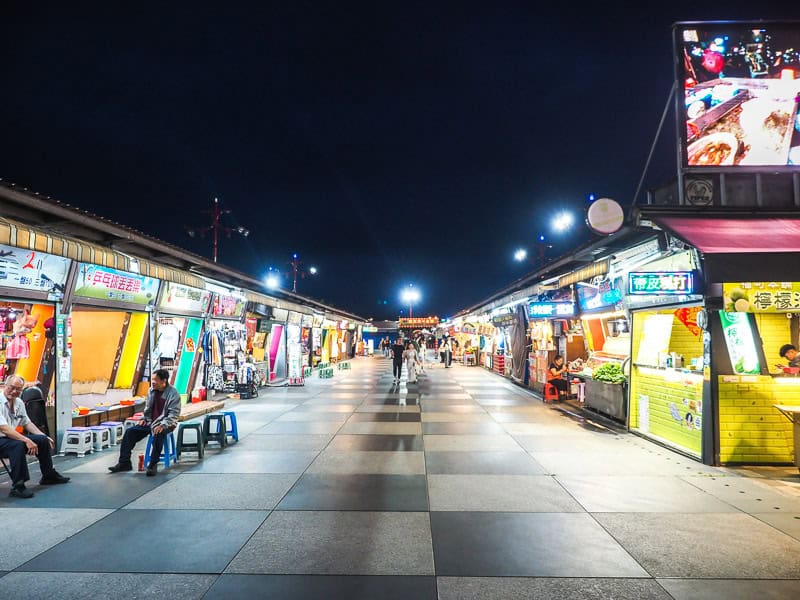 Looking up a night market street with lit up signs and food stalls along either but almost totally empty except for a single couple walking