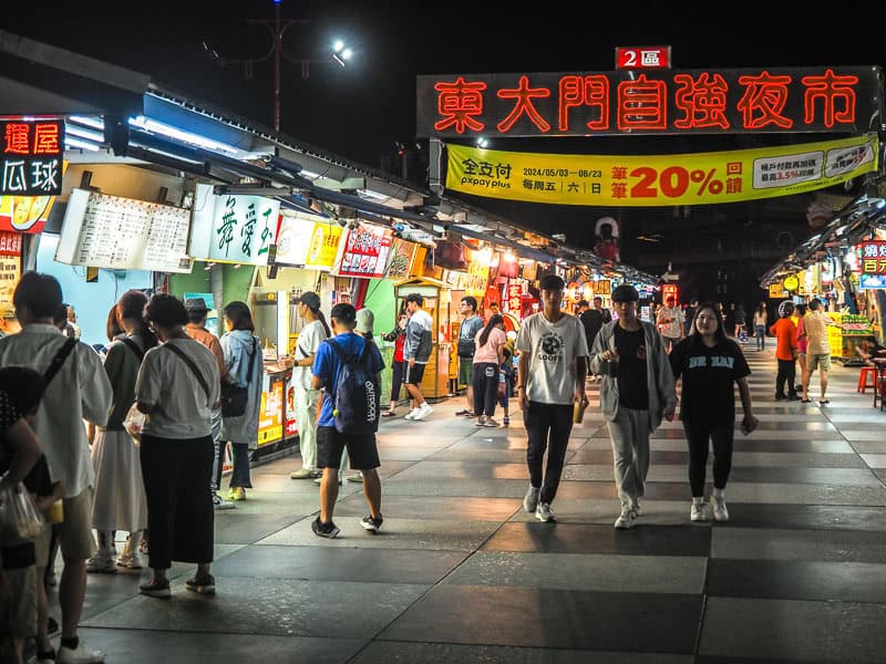 Some crowds of people walking in a dark night market with some stalls on the side with bright lights and a sign above with the market's name on neon red Mandarin characters