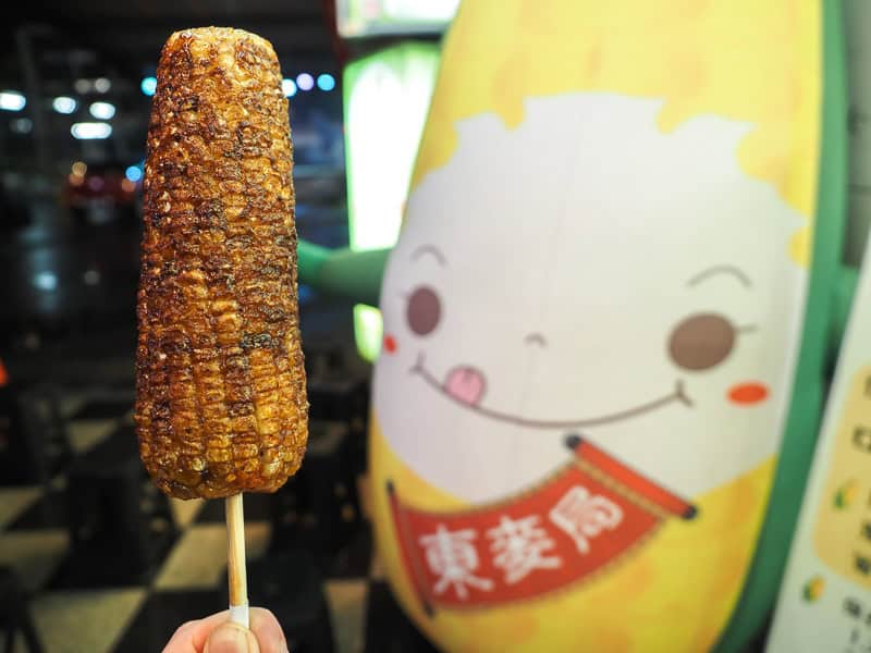 A hand holding up a stick of grilled corn on the left with a tall corn mascot with smiling face behind on the right