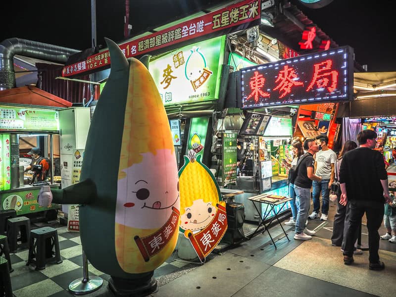 A night market vendor with tall corn statue beside it and some customers waiting for food on the right
