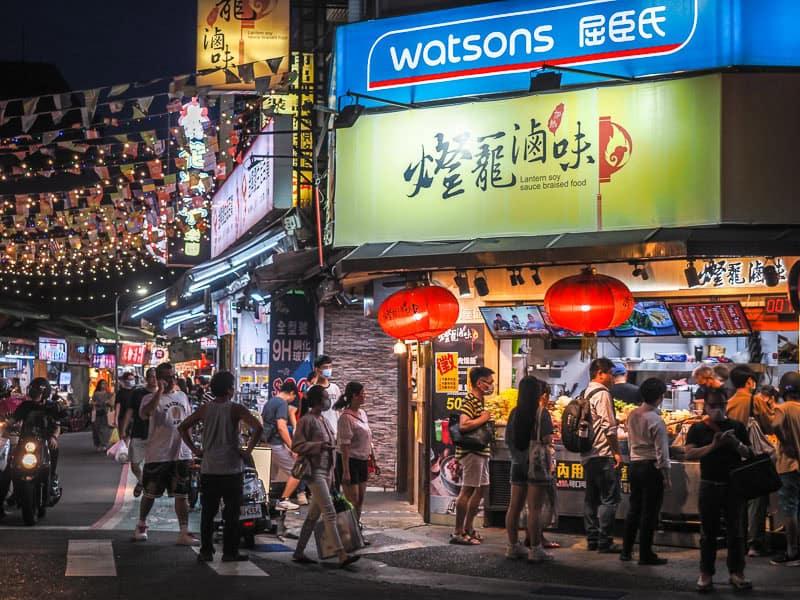 A street corner with a popular braised foods stall with yellow sign and two big red hanging lanterns and large blue sign for Watsons above, shot at night, with lots of people standing around waiting for food