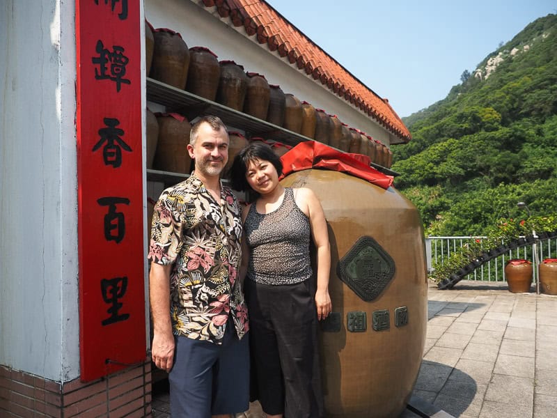 Nick Kembel standing with his wife next to a huge vessel of liquor in front of Matsu Liquor Factory