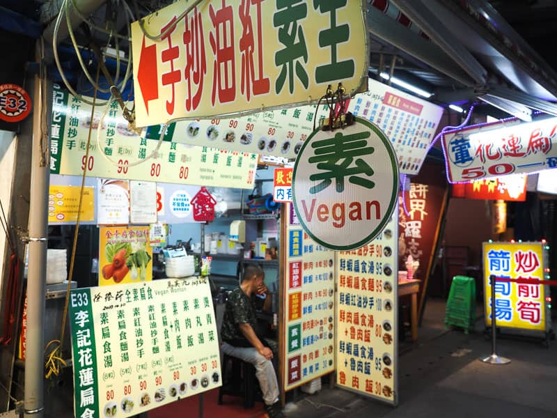 A night market restaurant front with many white signs with green and red Mandarin characters and the English word "vegan" pointing in