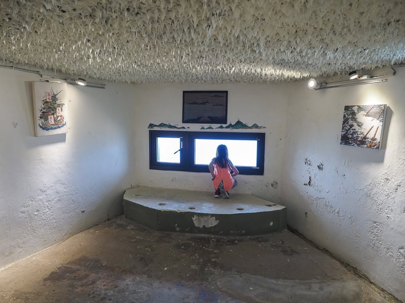 A young girl looks out a thin, horizontal window inside an empty room in a fort painted all in white