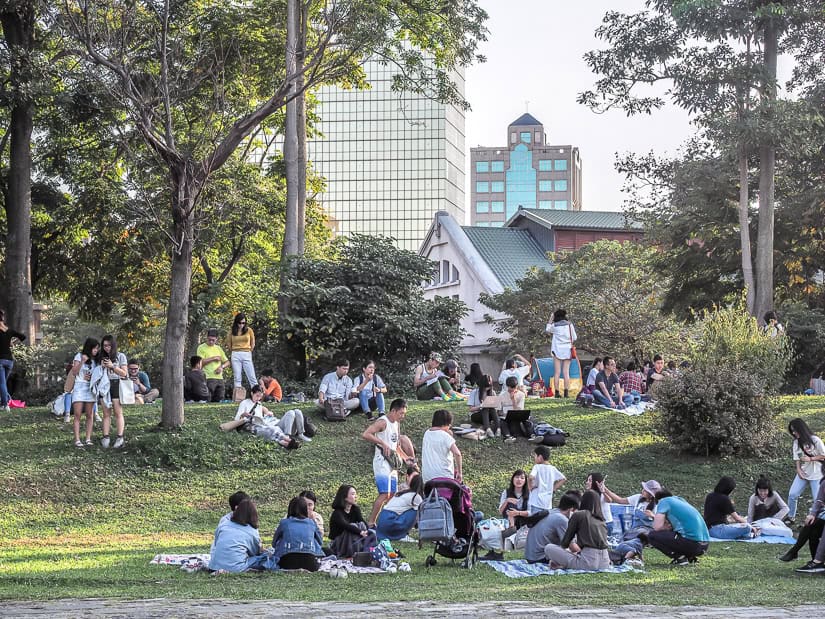 A grassy lawn with groups of Taiwanese having a picnic and some historic buildings behind the trees