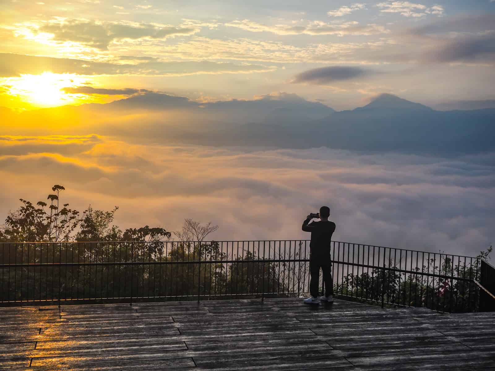 A person shot from behind on a large wooden platform overlooking a sunrise over a sea of clouds