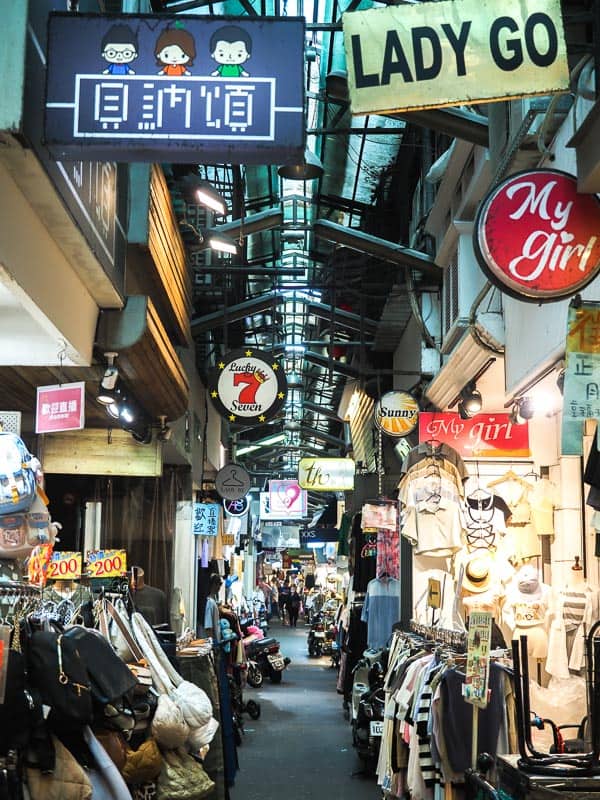 Looking down an aisle in a covered clothing market with many small cheap clothing shops on either side