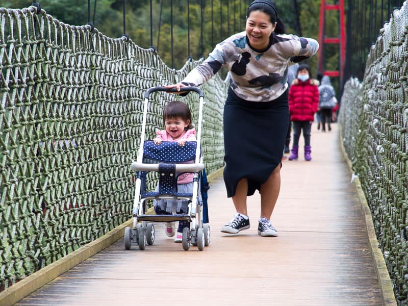 A Taiwanese woman pushing a stroller along a narrow suspension bridge, with her toddler aged daughter helping to push it along, and both are laughing