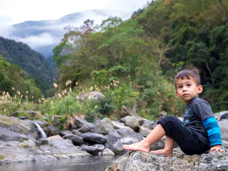 A young boy sitting on a rock beside a river with small waterfall and mountains behind