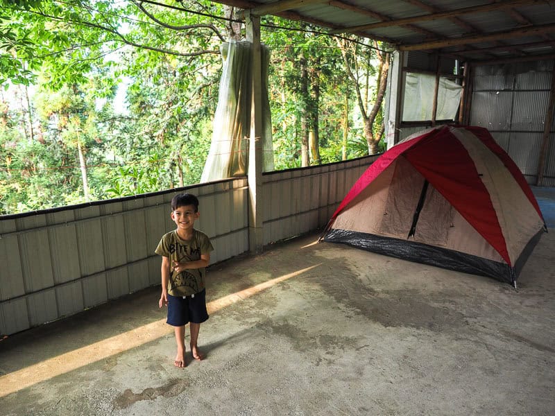 A young boy standing with no shoes on a cement floor in front of a tent with canopy above and forest outside