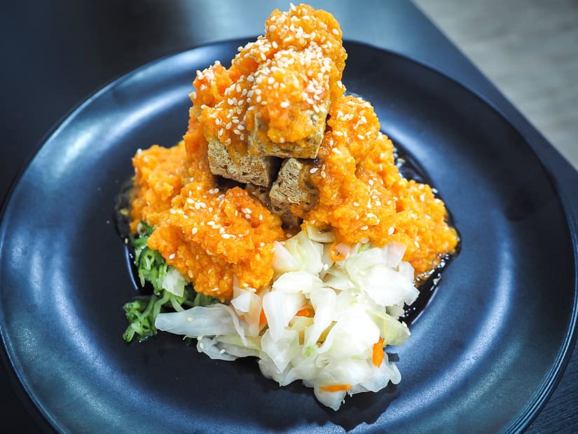 A black plate with a mound of stinky tofu cubes topped with orange crab roe and pickled vegetables on the side