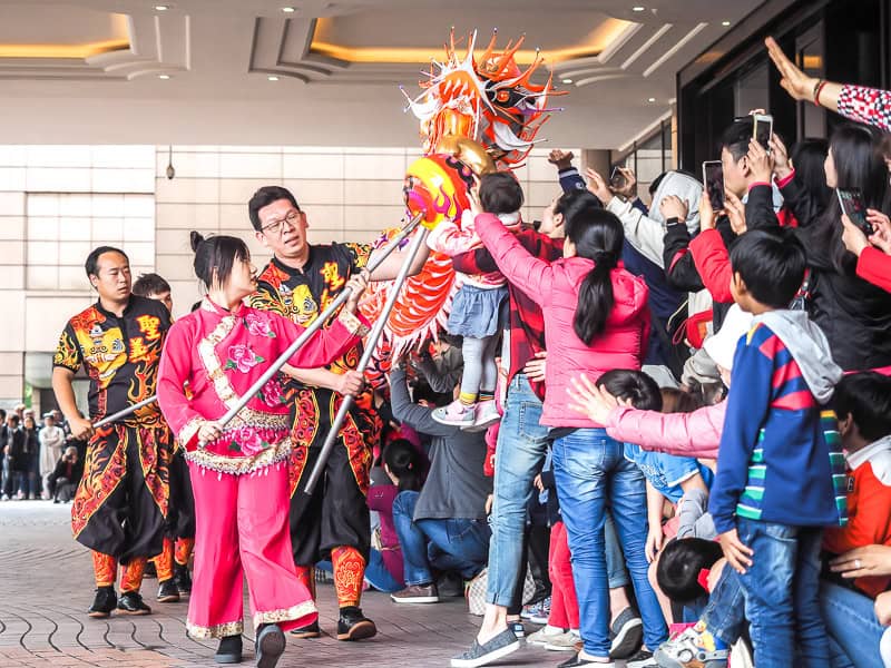 A crowd of children watch and try to touch a dragon as performers walk it past them