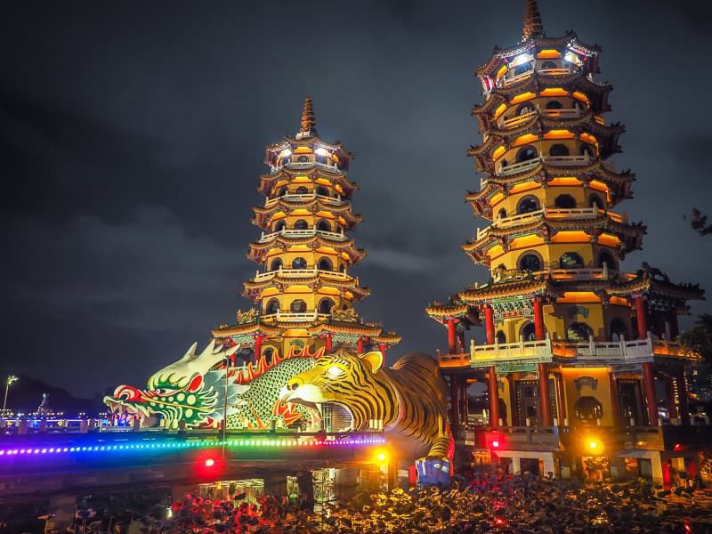 Two pagodas at night, with a tiger and dragon statue at the bottom of each and colorful lights on them