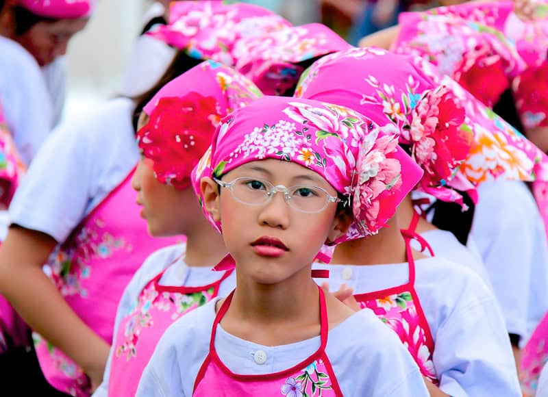 Some Hakka kids dressed in pink, including a boy with glasses and make up looking directly at the camera
