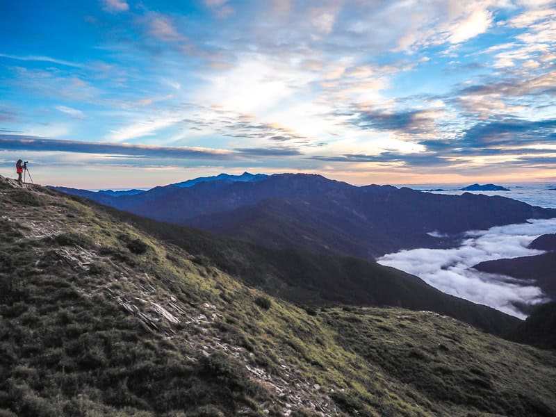 A large valley filled with a a sea of clouds at sunrise, with one person standing on a mountain-top on the left photographing it