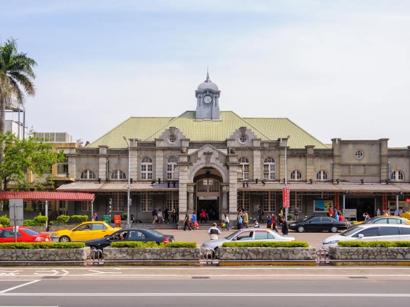 The exterior of a large historic train station with line of cars and taxis out front, shot from across the street