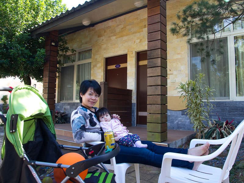 A young Taiwanese mother relaxing with feet up on a white plastic chair and holding a baby in front of a cabin