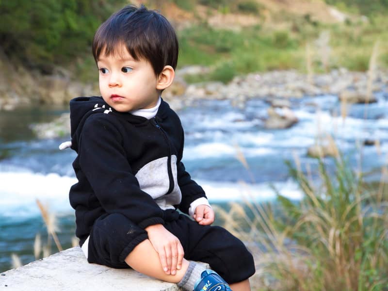 A young boy in black onesie sitting on a cement gate with river flowing behind him