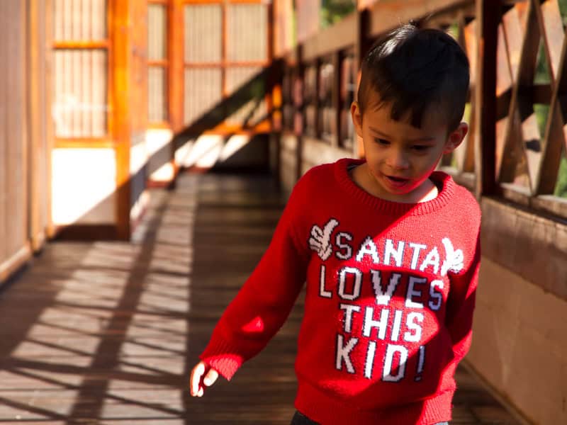 A young boy with red sweater that says "Santa Loves this Kid" walking on a wooden, sun-lit balcony