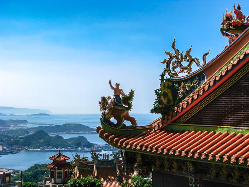 A temple's orange roof and statues on the right, with a view down to the coast on the left 