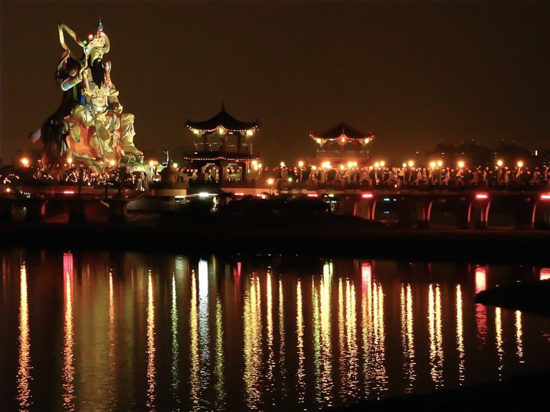 A walkway leads across water to a huge Taoist god statue at night