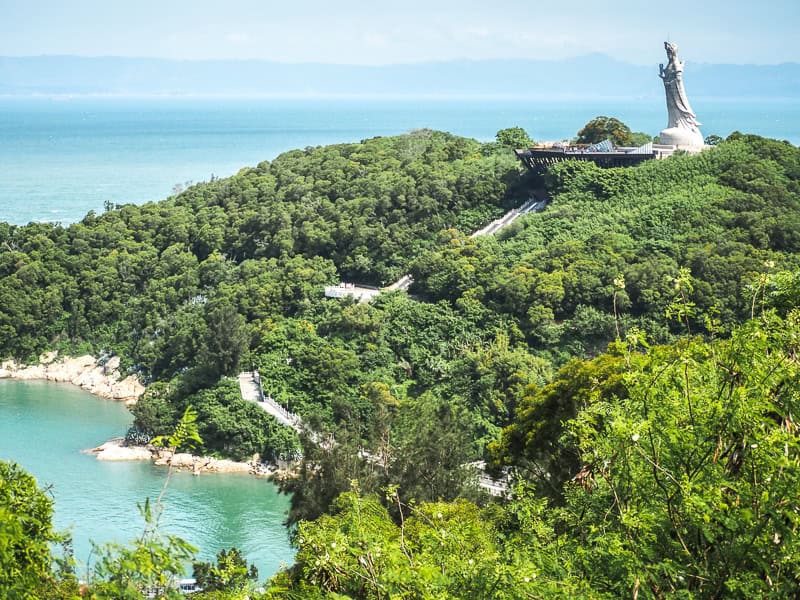 A large Matsu statue atop of a coastal hill, with stairs leading up to it, and a mainland visible beyond the sea.