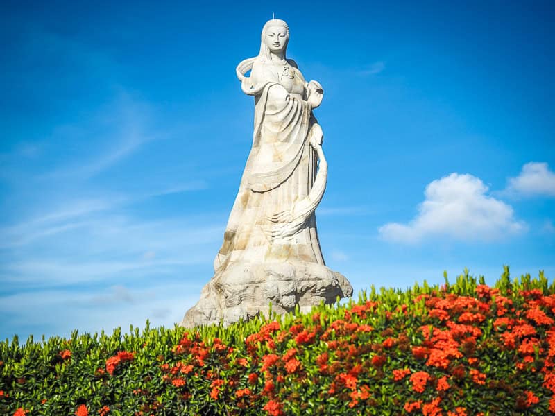 A tall white statue of Matsu with bed of red flowers at the bottom