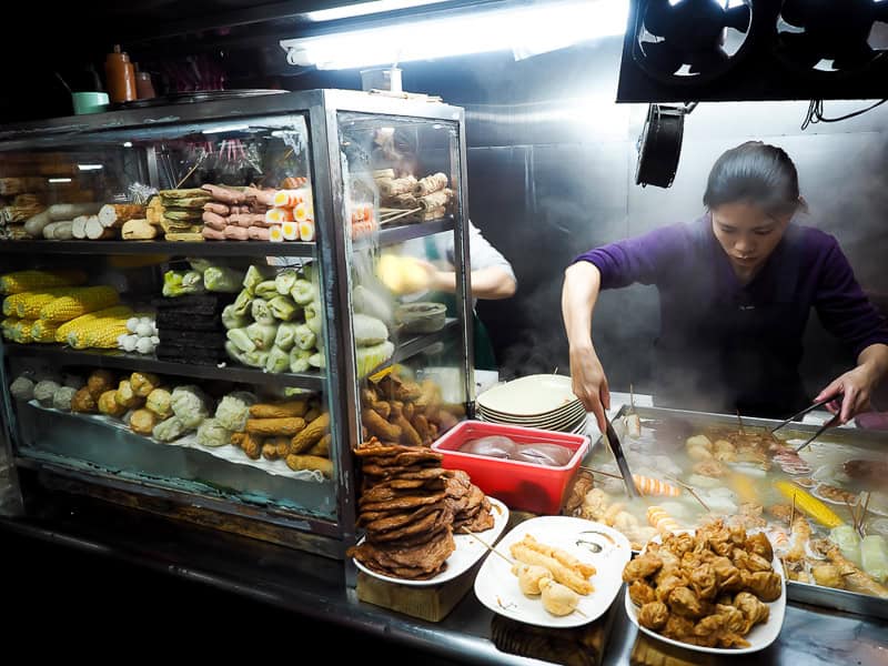 A female vendor tends to some foods in steaming vats in a night market stall